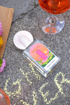 Shifumi Eau de Parfum – klares Glasflakon mit farbenfrohem Etikett, rosafarbener Deckel, grauer Hintergrund – Parfüm