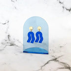 Spectral Blue Earrings – blue acrylic shape with gold setting, on blue acrylic stand, marble background – jewelry