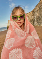 Shells Organic Cotton Badetuch – rosafarben mit weißen Muschelmustern, am Strand getragen – Strandaccessoires