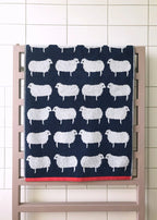 Sheep Organic Cotton Badelaken – dunkelblaues Strandtuch mit weißen Schafen, roter Saumkante, auf Holzständer – Badetuch