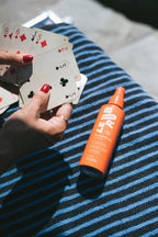 Kartenspel am Strand, gut geschützt mit Sunscreen Mist SPF 50 Sprühflasche auf blau-weißem Handtuch, Sonnenschutz für den Körper.