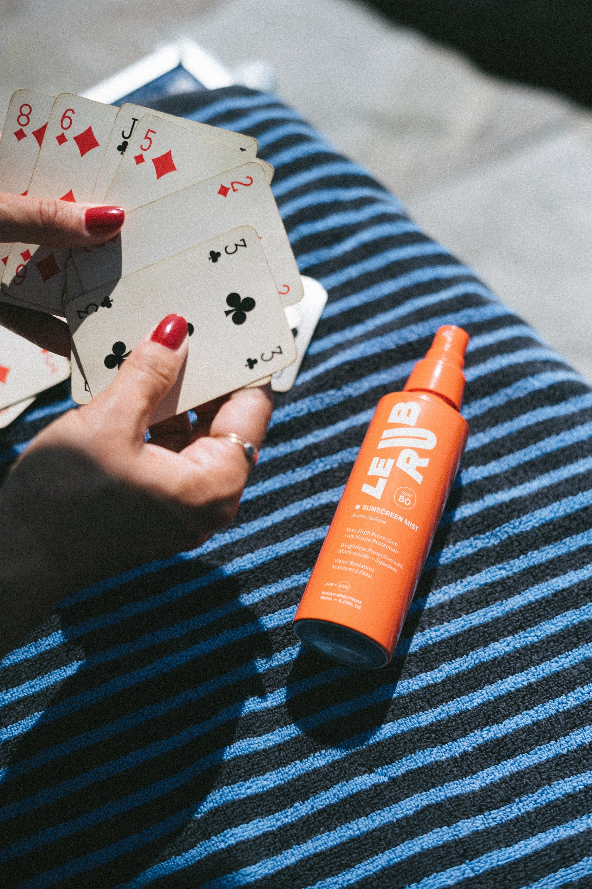 Kartenspel am Strand, gut geschützt mit Sunscreen Mist SPF 50 Sprühflasche auf blau-weißem Handtuch, Sonnenschutz für den Körper.