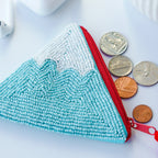Small Mountain Clutch, eine kleine, dreieckige Perlenhandtasche in Blau und Weiß mit rotem Reißverschluss und Münzen daneben.