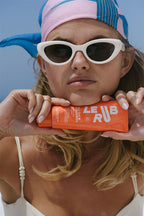 Frau mit Sonnenbrillen und Le Rub Everyday Sunscreen SPF50 in einer orangefarbenen Tube vor blauem Hintergrund, Hautpflege.
