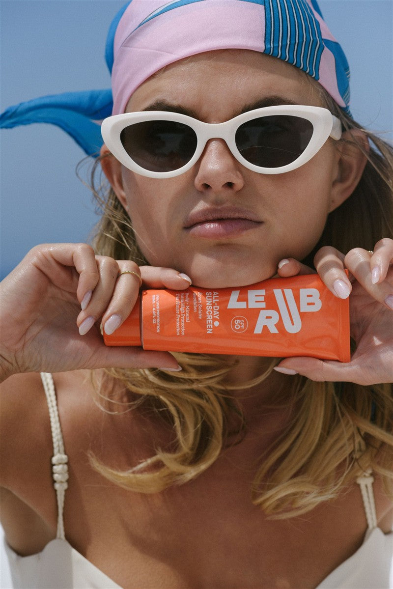 Frau mit Sonnenbrillen und Le Rub Everyday Sunscreen SPF50 in einer orangefarbenen Tube vor blauem Hintergrund, Hautpflege.