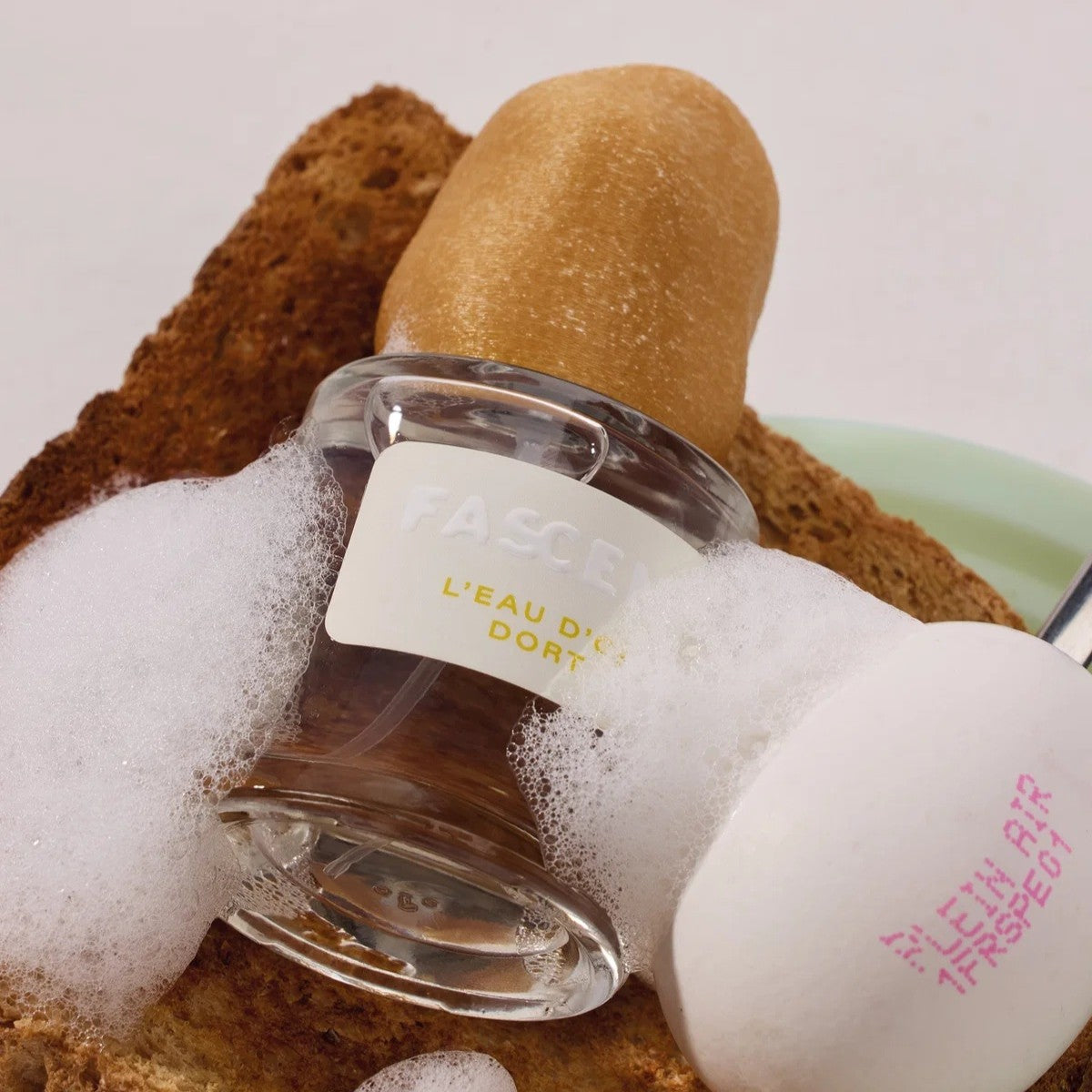 Fascinate L'eau D'or Dort Eau de Parfum – klares Glasfläschchen mit weißem Etikett, umgeben von Schaum – Parfüm