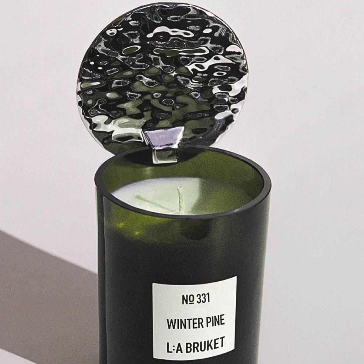 L:A Bruket Candle Reflektor, grünes Glasgefäß mit silbernem Metalldeckel vor hellem Hintergrund, Duftkerze.