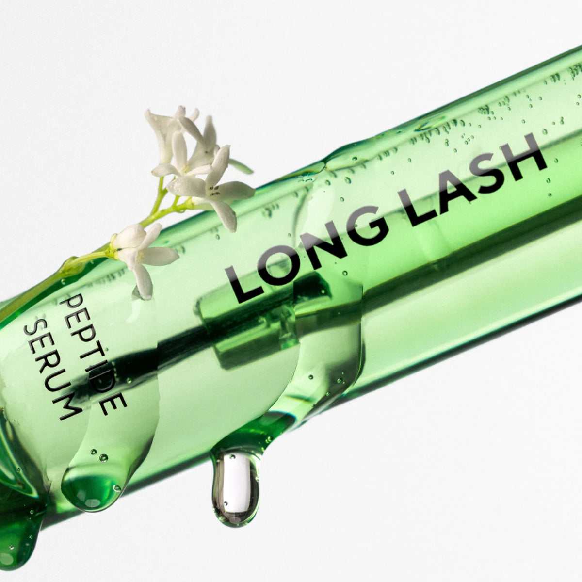 GreenGlam Long Lash Peptid-Wimpernserum: Nahaufnahme einer grünen Flasche mit Serum-Tropfen und kleinen weißen Blüten.