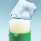 SHOT Koffein-Augenserum-Stick: Ein Stück Eis liegt auf dem ausgefahrenen, cremefarbenen Serum-Stick in einem grünen Gehäuse.