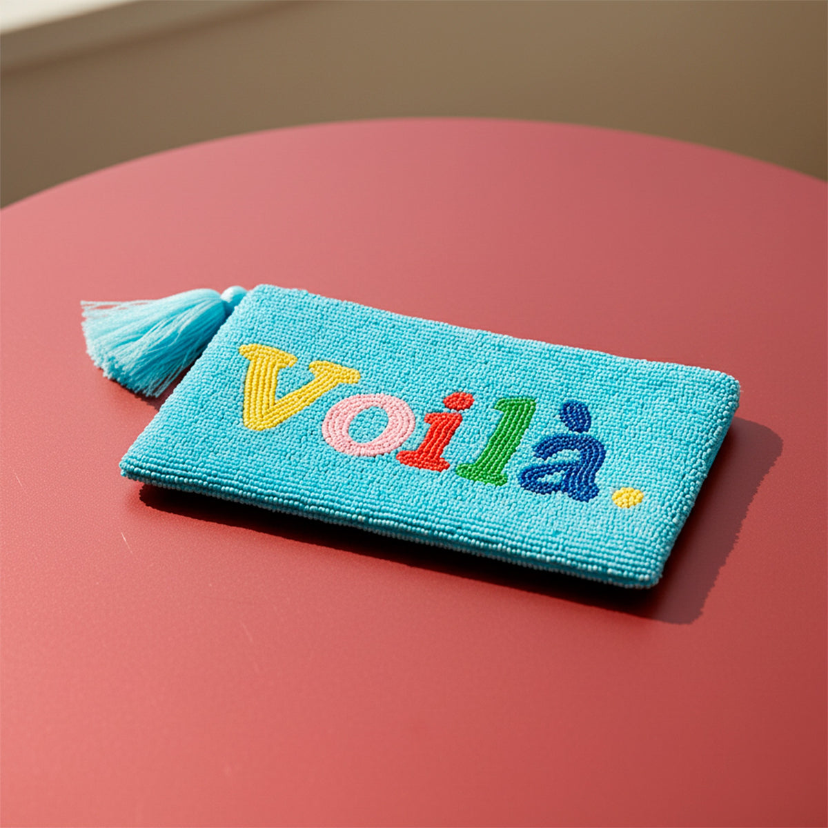 Voilà Clutch, hellblaue, perlenbestickte Clutch mit farbigem Schriftzug auf rötlichem Hintergrund.